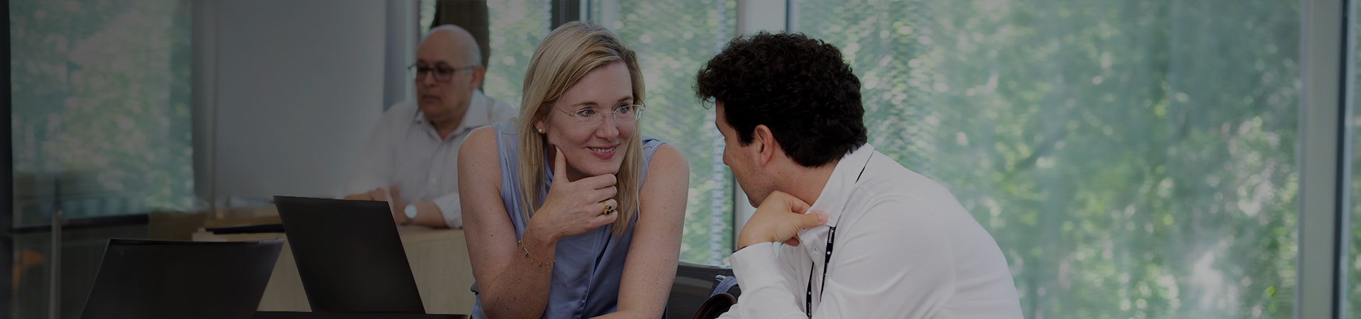 3 people sitting, one in the background and two, a women and a man in the foreground. The two in the foreground are talking and looking at each other and the women is smiling